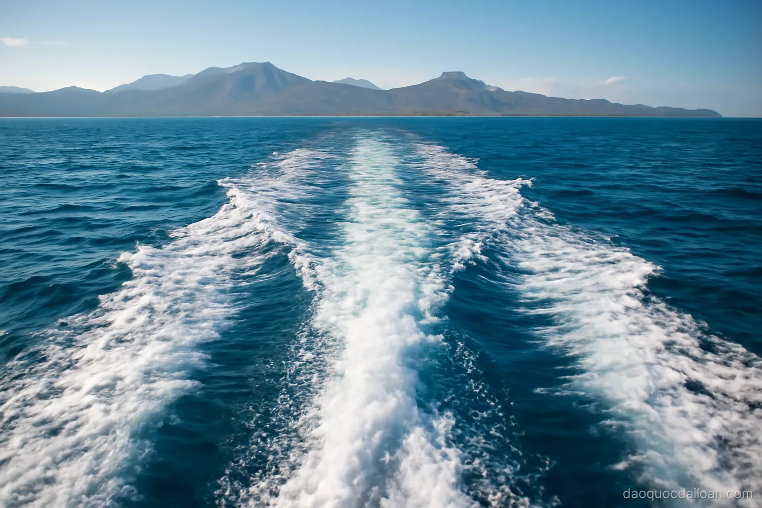 Deep blue open ocean water with a boat wake and distant mountainous coastline, bright daylight, no readable text