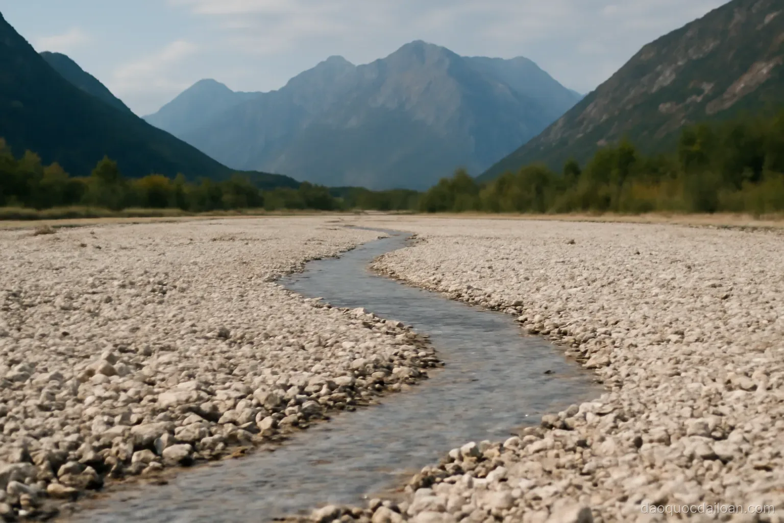 A wide dry riverbed of pale gravel with a thin stream winding through it, mountains behind, no readable text